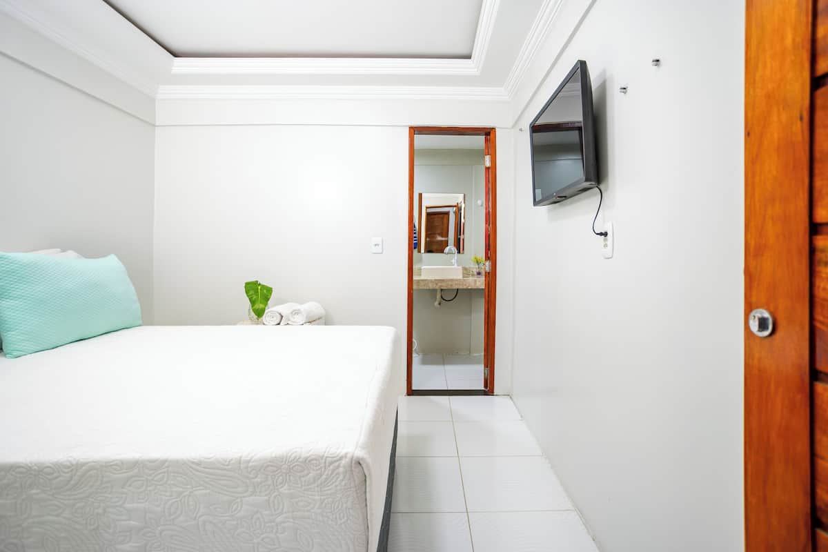 Une chambre à coucher avec un lit, un téléviseur fixé au mur et une salle de bain visible à travers une porte ouverte.