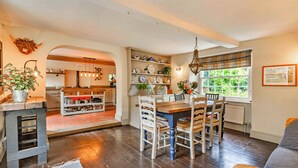 A dining area with a wooden table and chairs, a kitchen in the background, and a window with a view of greenery.