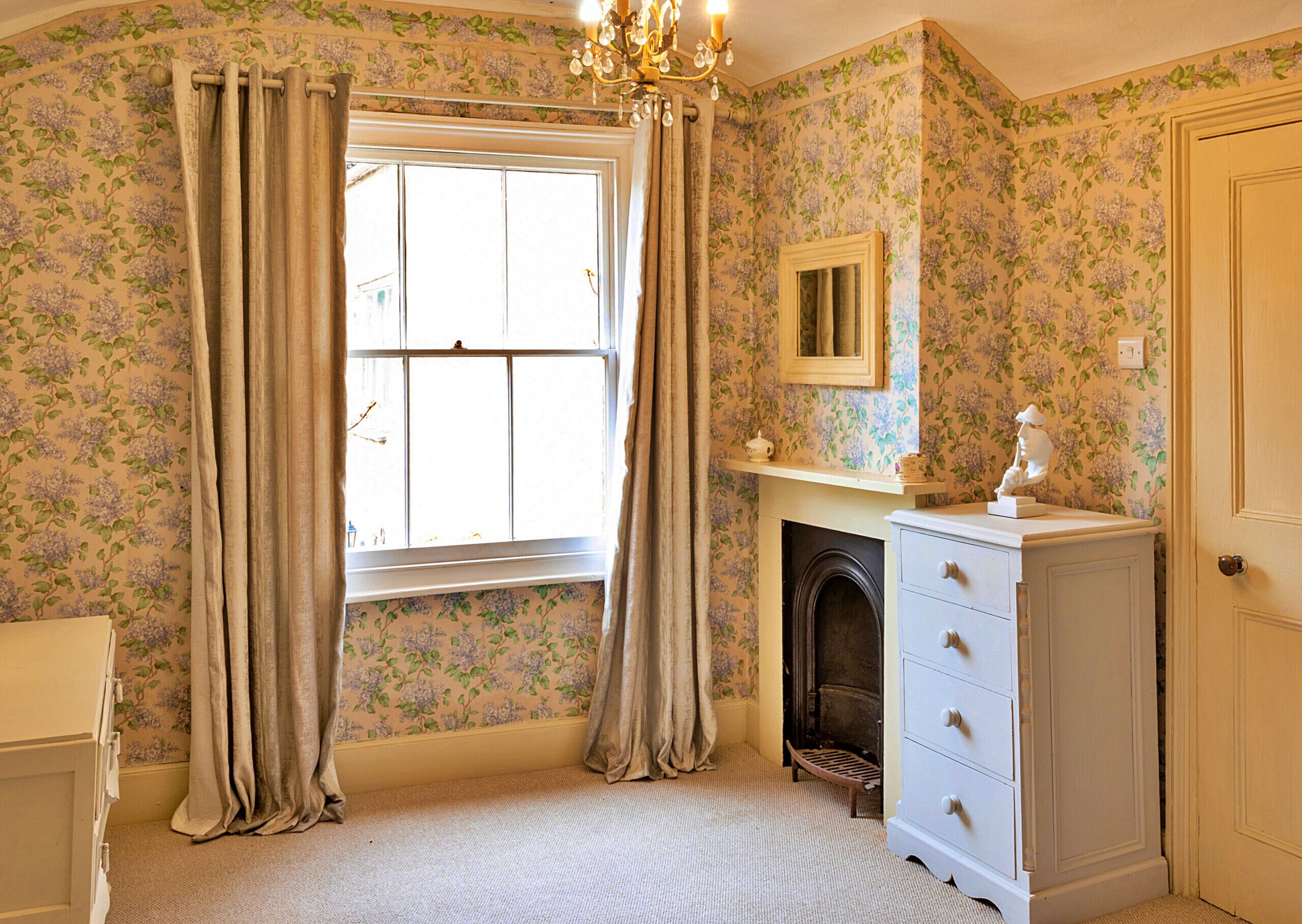 A room with floral wallpaper, a fireplace, a dresser, and a window with curtains.