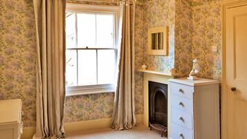 A room with floral wallpaper, a fireplace, a dresser, and a window with curtains.