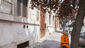 A narrow street with a parked orange scooter, a tree, and a building with a wooden door and shutters. - femasuites (Rome)