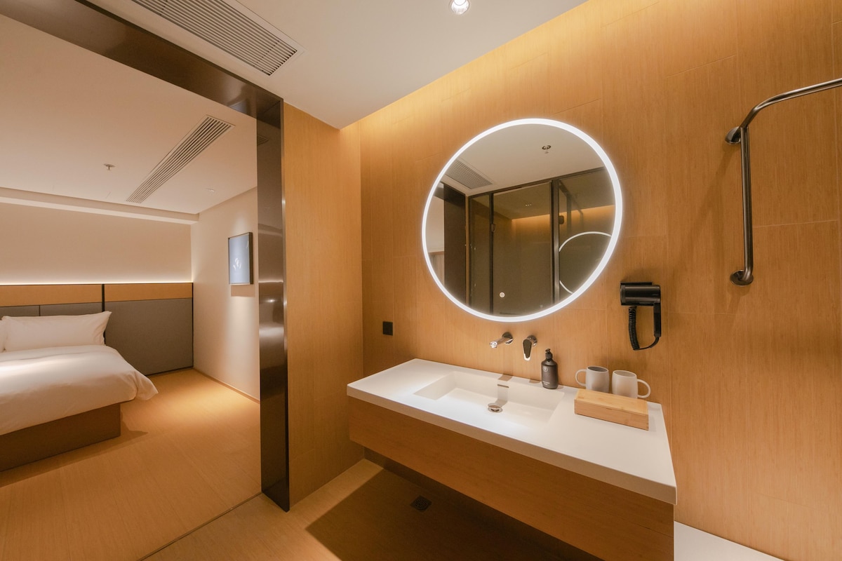 A modern hotel room with a bed, a large round mirror, a white sink, and a handheld shower.
