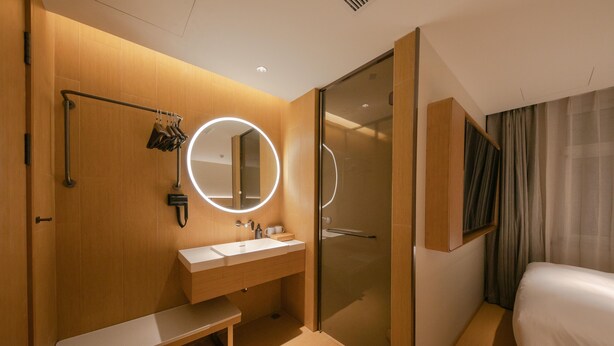 A modern hotel bathroom with a wooden vanity, a round mirror with lights, a shower, and a bed. - Ji Hotel (Beijing)
