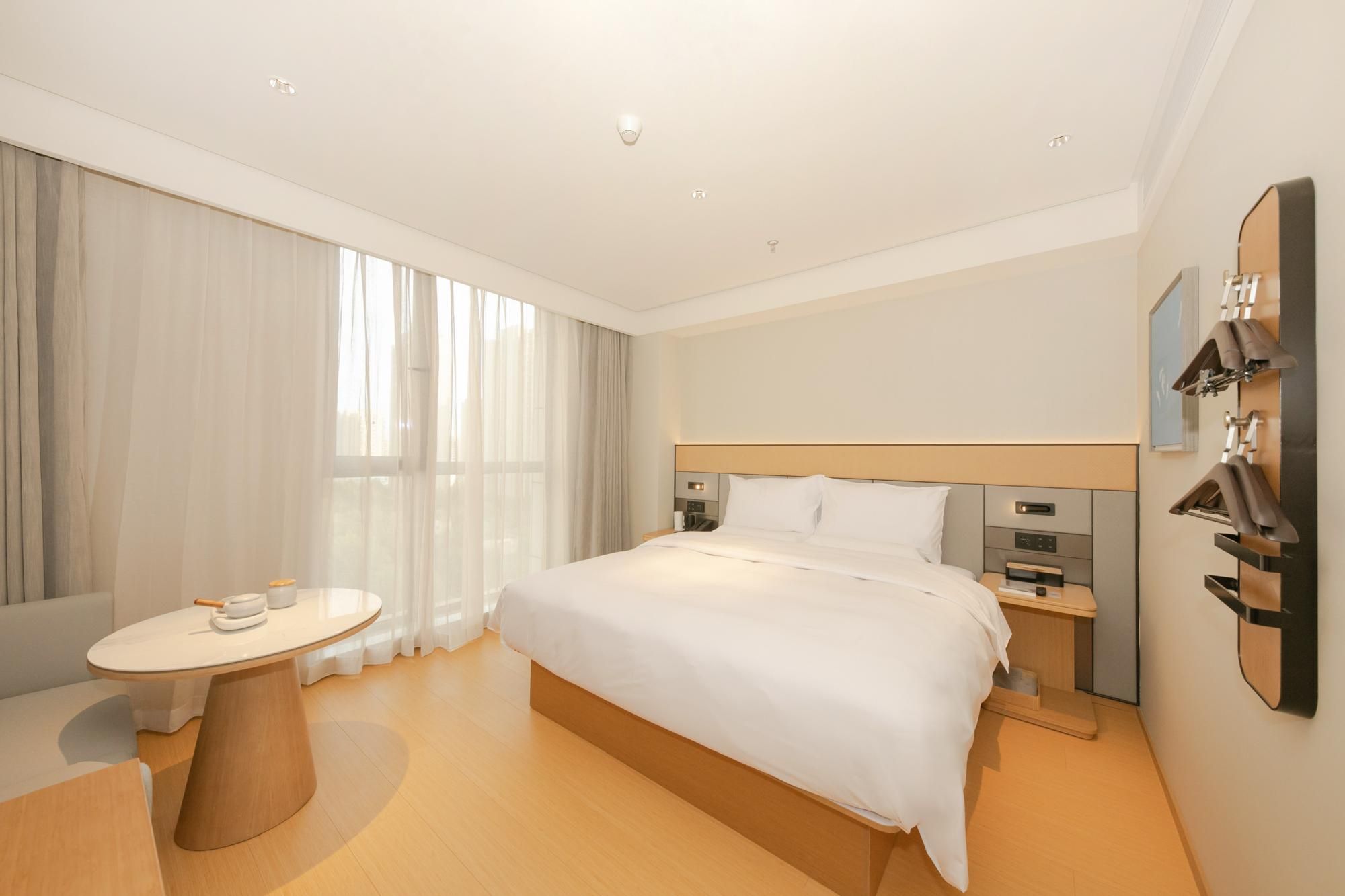 Una habitación de hotel moderna con una cama grande, mesitas de noche, una pequeña mesa redonda con una silla y un amplio ventanal con cortinas.