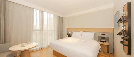 Una habitación de hotel moderna con una cama grande, mesitas de noche, una pequeña mesa redonda con una silla y un amplio ventanal con cortinas.