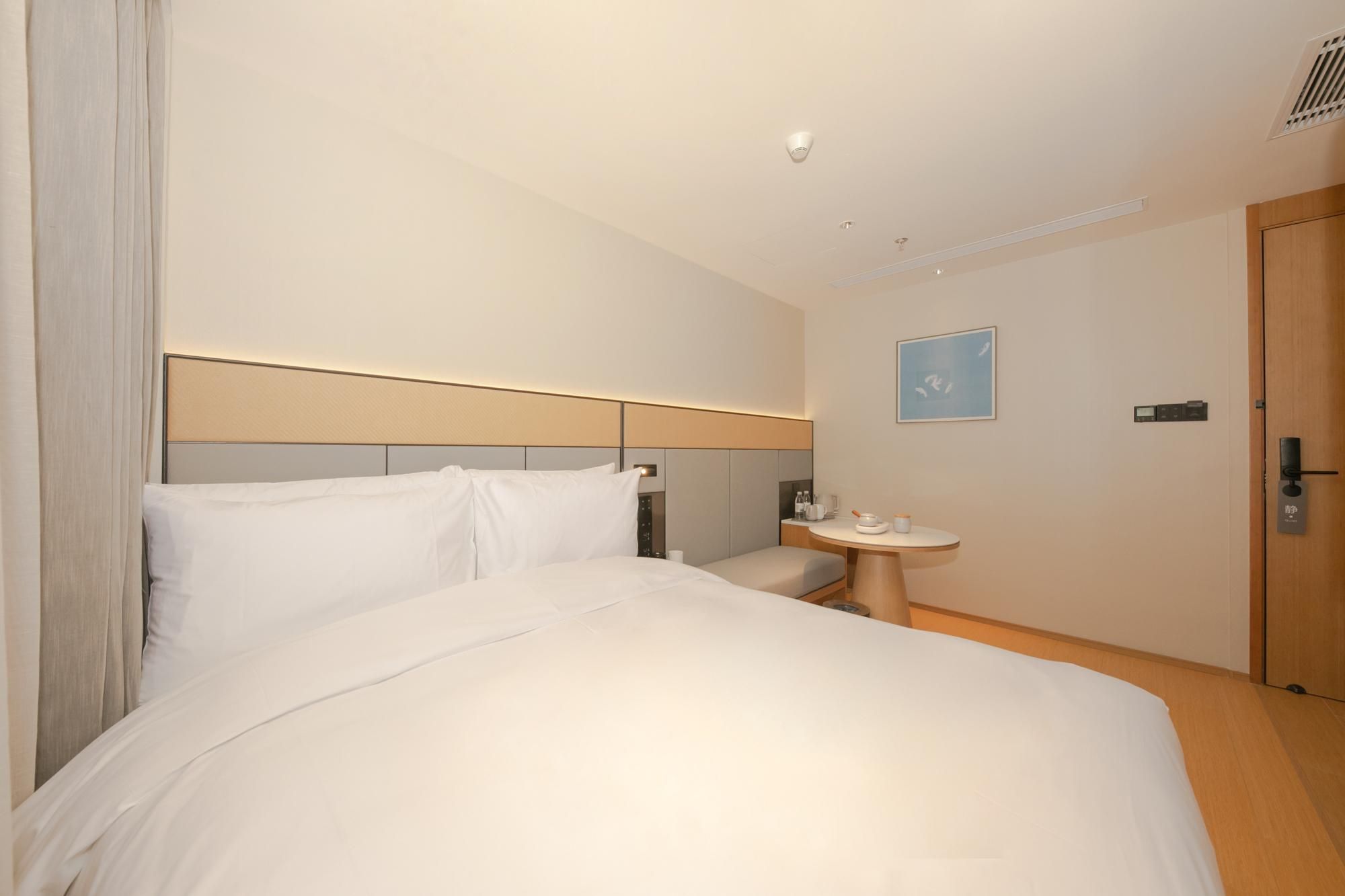 A hotel room with a large bed, a bedside table with a lamp, a small round table with a cup and saucer, and a door with a lock.