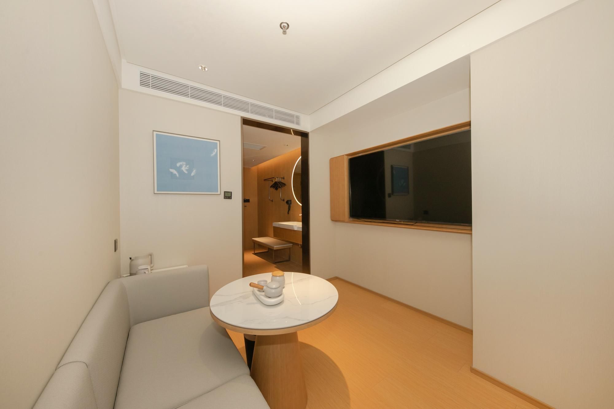 Ett modernt hotellrum med ett runt bord, en soffa, ett stort fönster och ett badrum som syns genom en öppen dörr.
