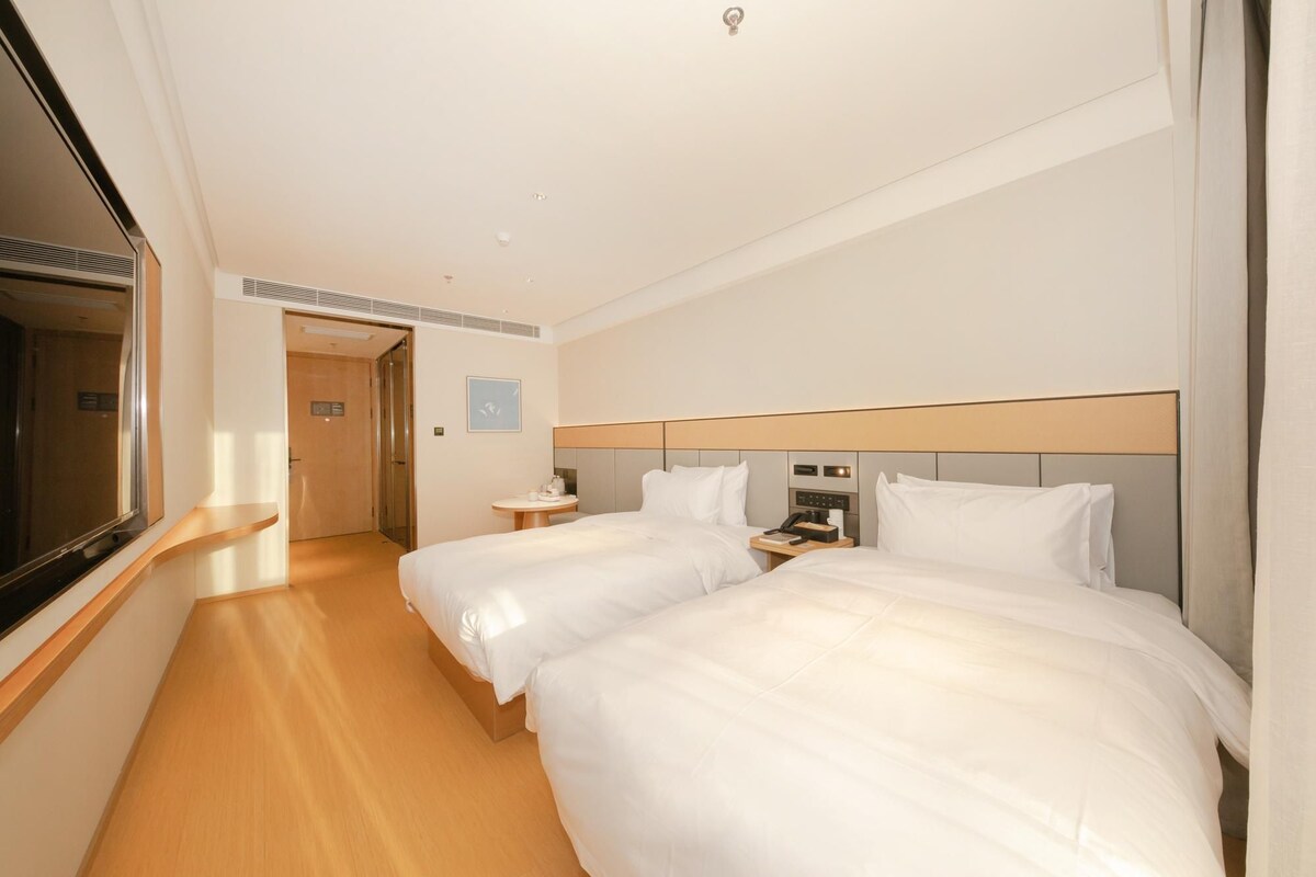 A hotel room with two beds, a bedside table with a phone and a lamp, a small round table with a chair, and a large window.