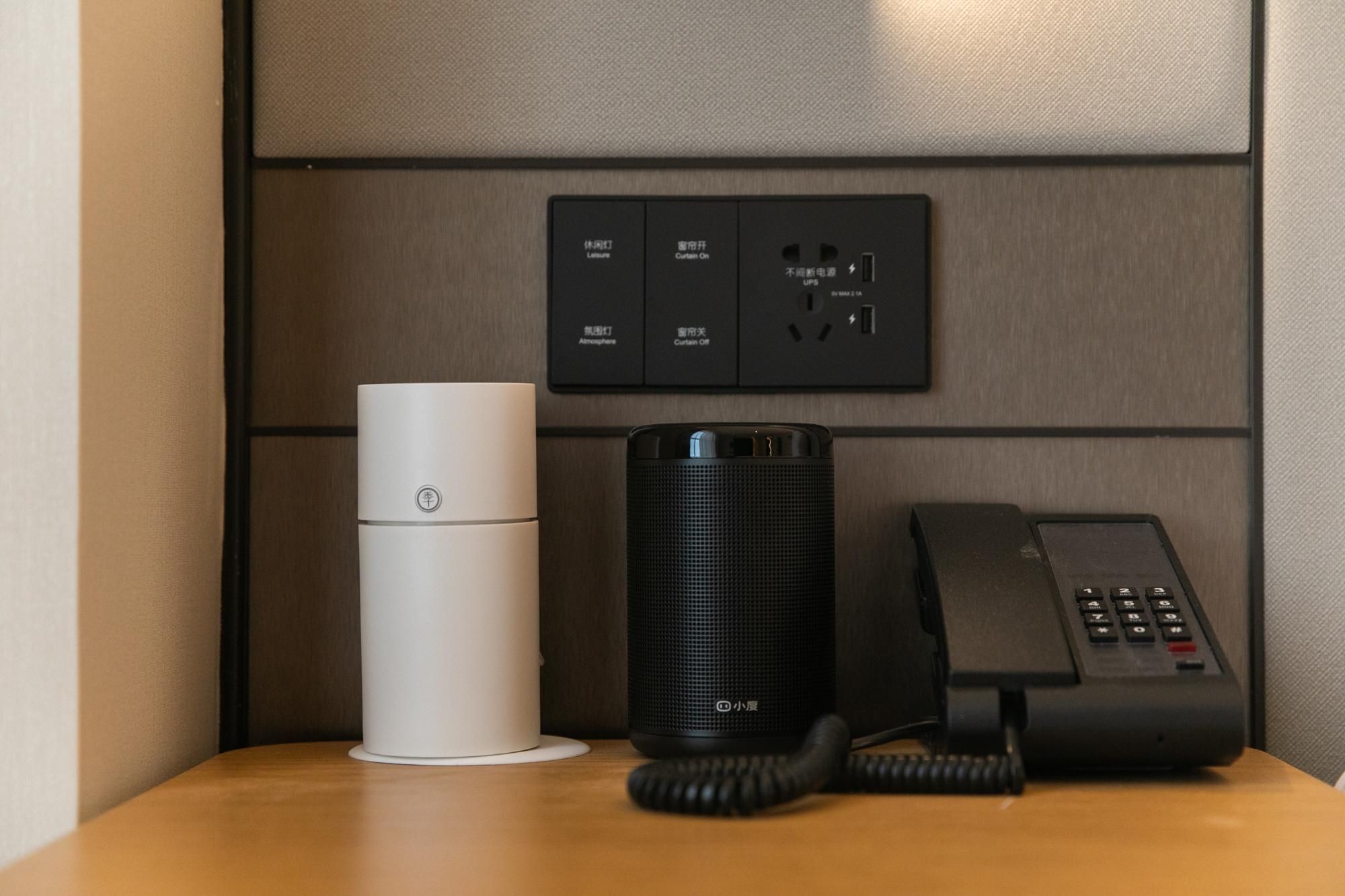 A hotel room with a white speaker, a black telephone, and a control panel with various settings.