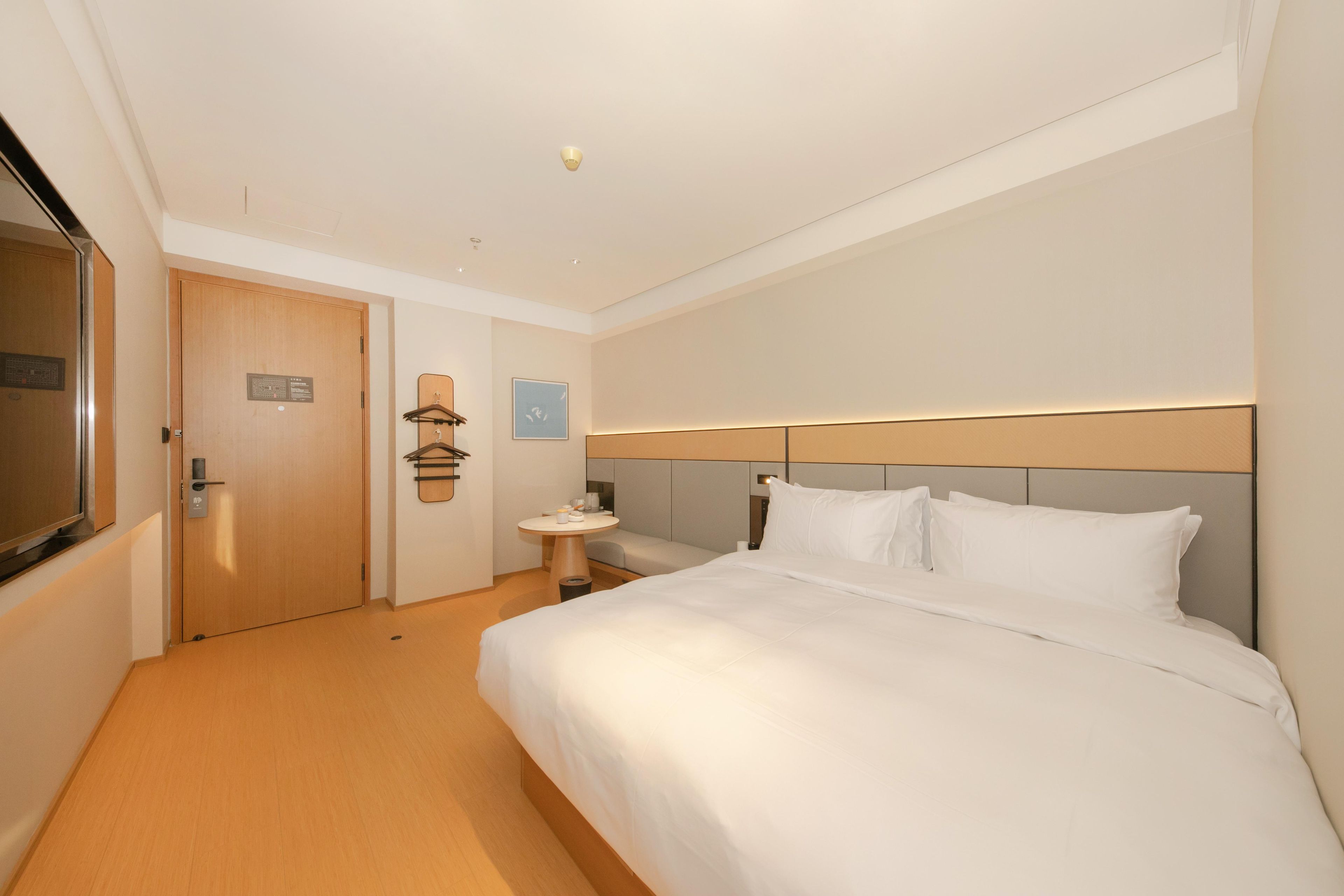 Habitación de hotel con una cama grande, una mesita de noche, una pequeña mesa redonda, un espejo y una puerta.