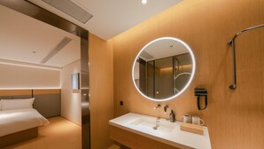 Une chambre d’hôtel moderne équipée d’un lit, d’un grand miroir rond, d’un lavabo blanc et d’une douchette à main.