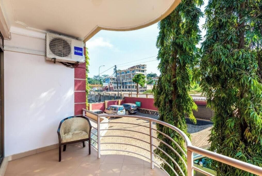 Een balkon met een stoel, een airconditioning en uitzicht op een gebouw en geparkeerde auto's.