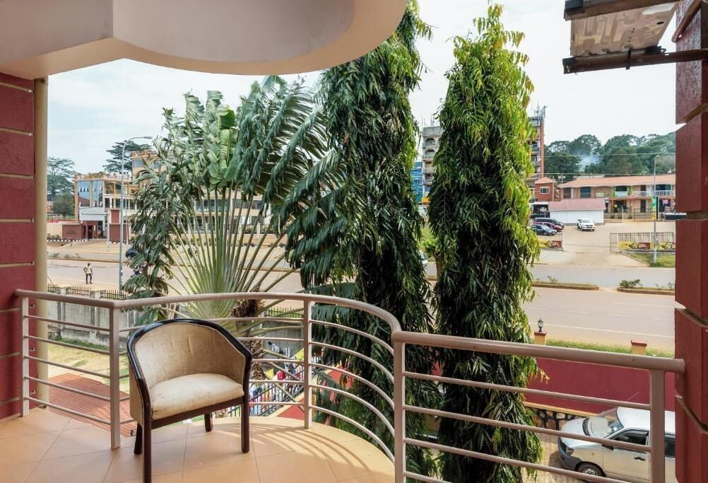 A balcony with a chair, surrounded by tropical plants, overlooking a street with buildings and parked cars.