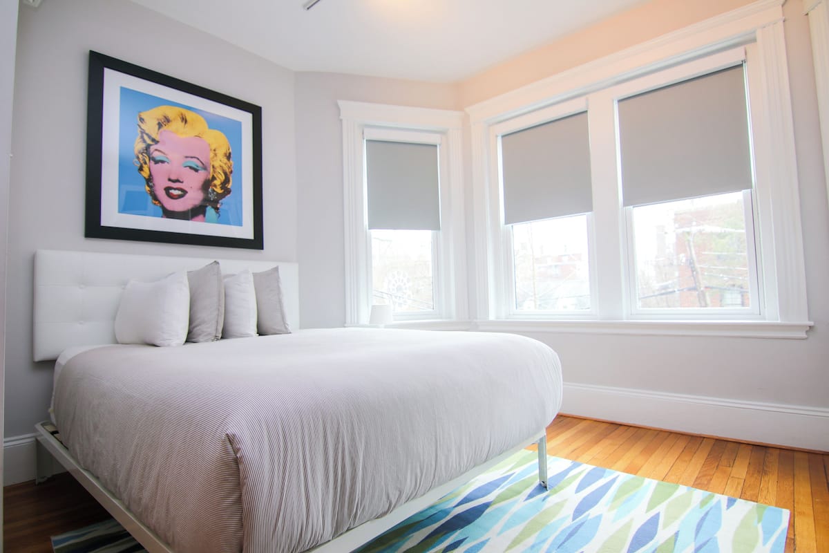 A bedroom with a bed, a framed artwork on the wall, and a window with blinds.