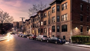 A row of brick townhouses with multiple stories, parked cars along the street, and a clear sky.