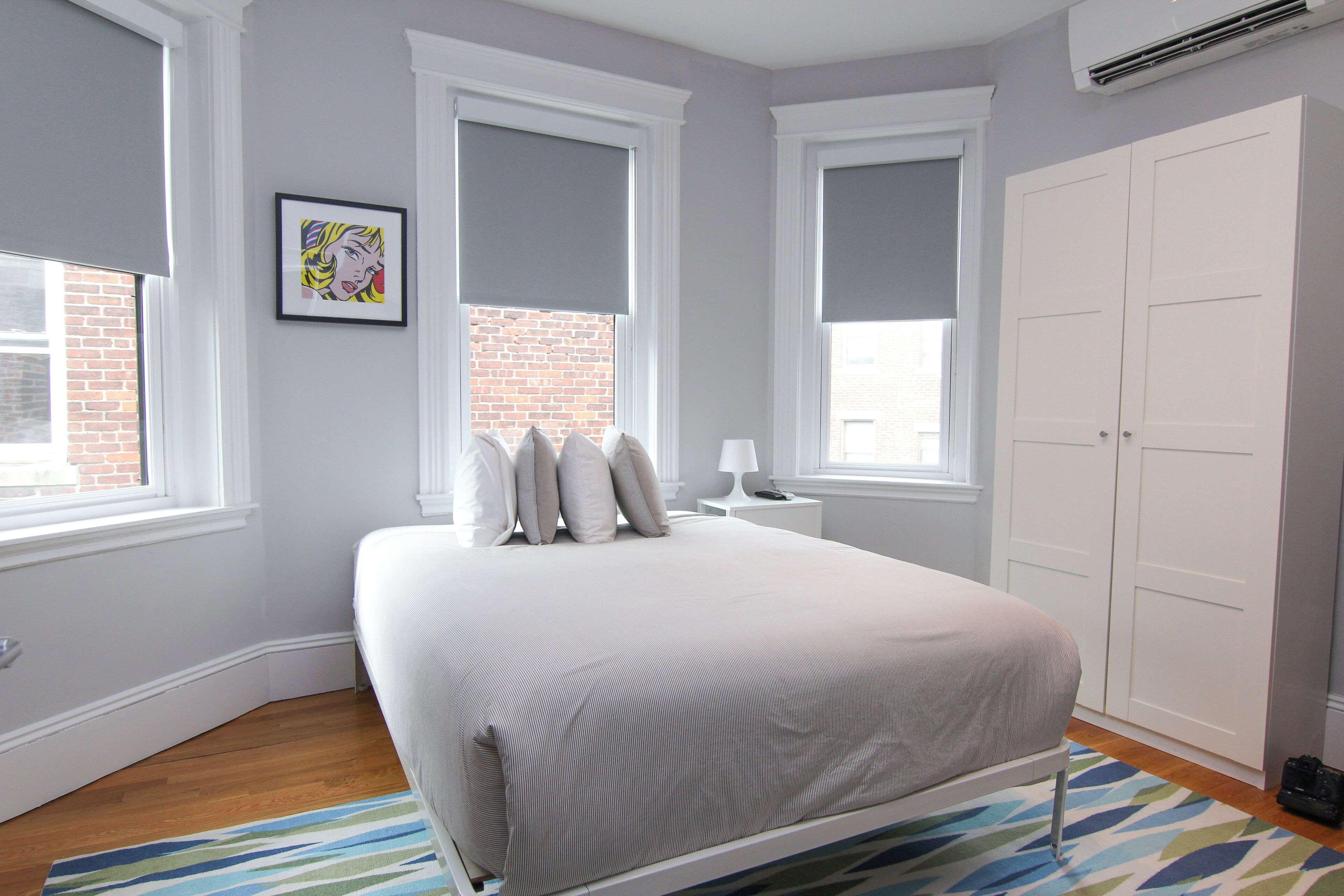 A bedroom with a bed, a nightstand, a lamp, a wardrobe, and a window with a view of a brick building.