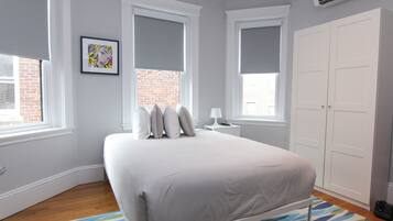 A bedroom with a bed, a nightstand, a lamp, a wardrobe, and a window with a view of a brick building.