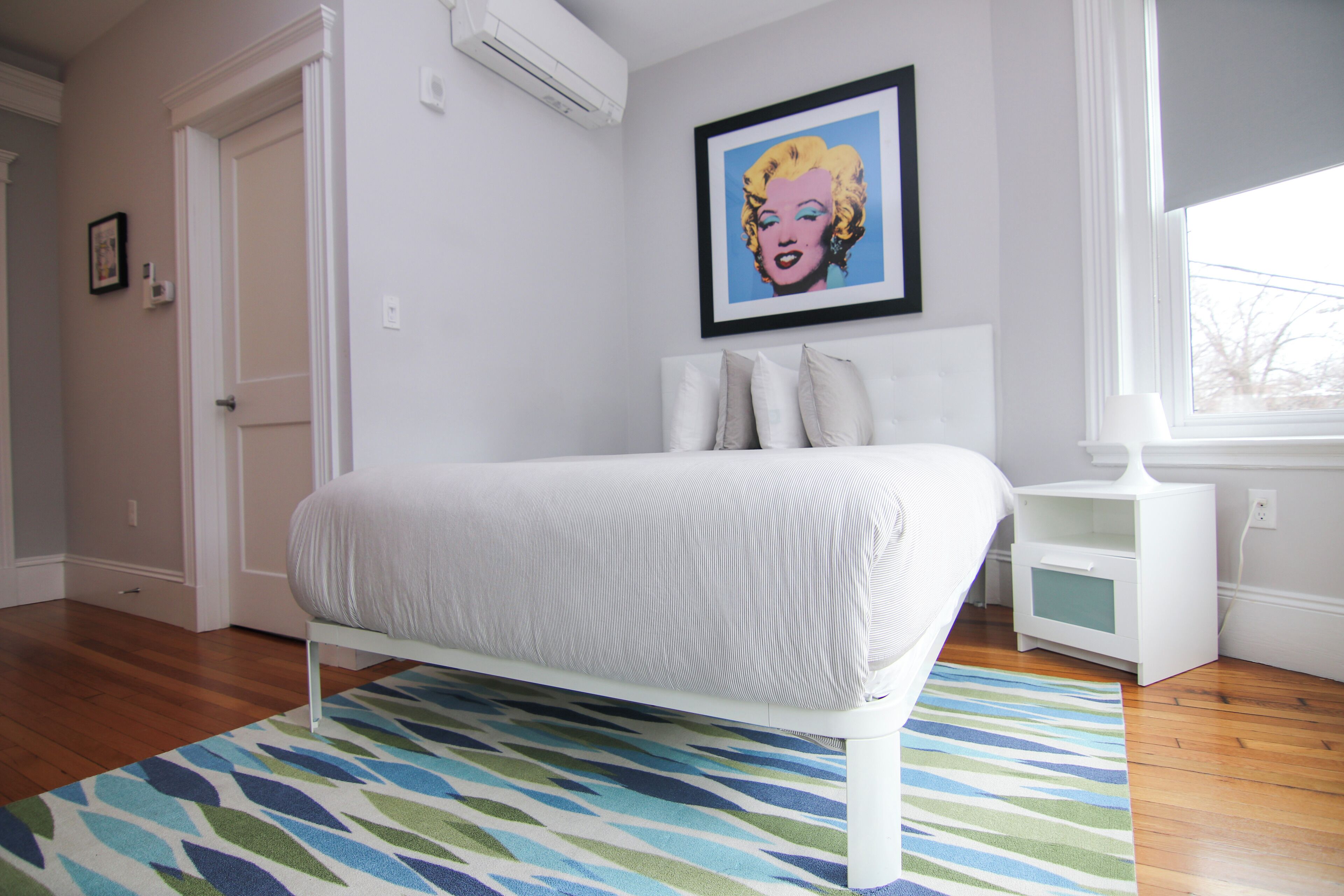 A bedroom with a bed, a patterned rug, a framed picture on the wall, and a nightstand.