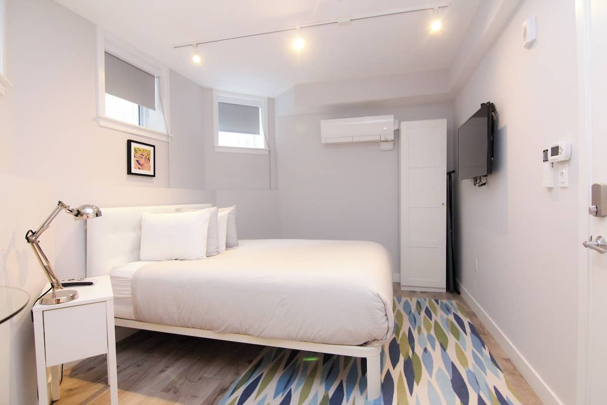A bedroom with a bed, a bedside table with a lamp, a window, an air conditioning unit, and a television mounted on the wall.
