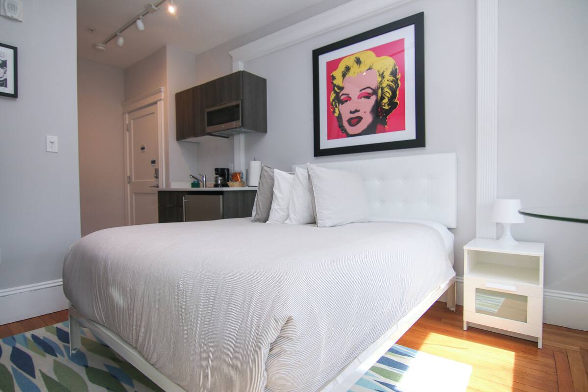 A bedroom with a large bed, a nightstand, a framed artwork on the wall, and a kitchenette in the background.