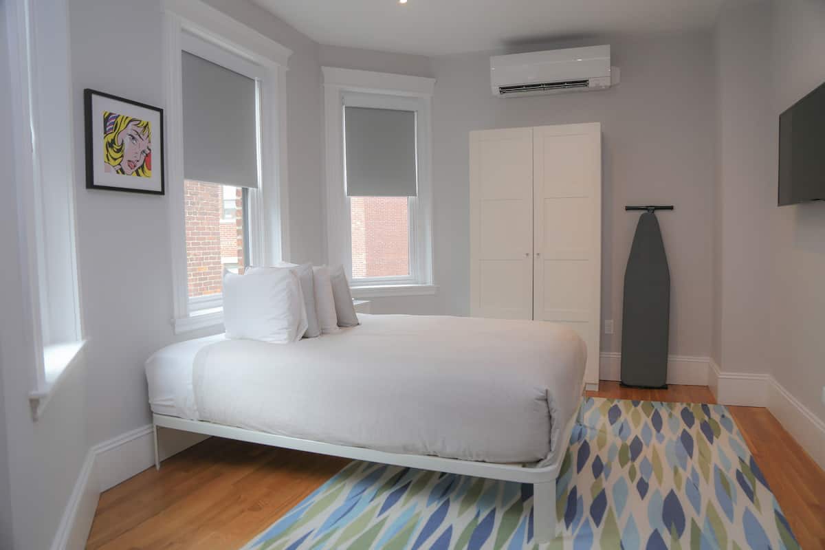 A bedroom with a bed, white walls, a window with blinds, a wardrobe, an ironing board, and an air conditioning unit.