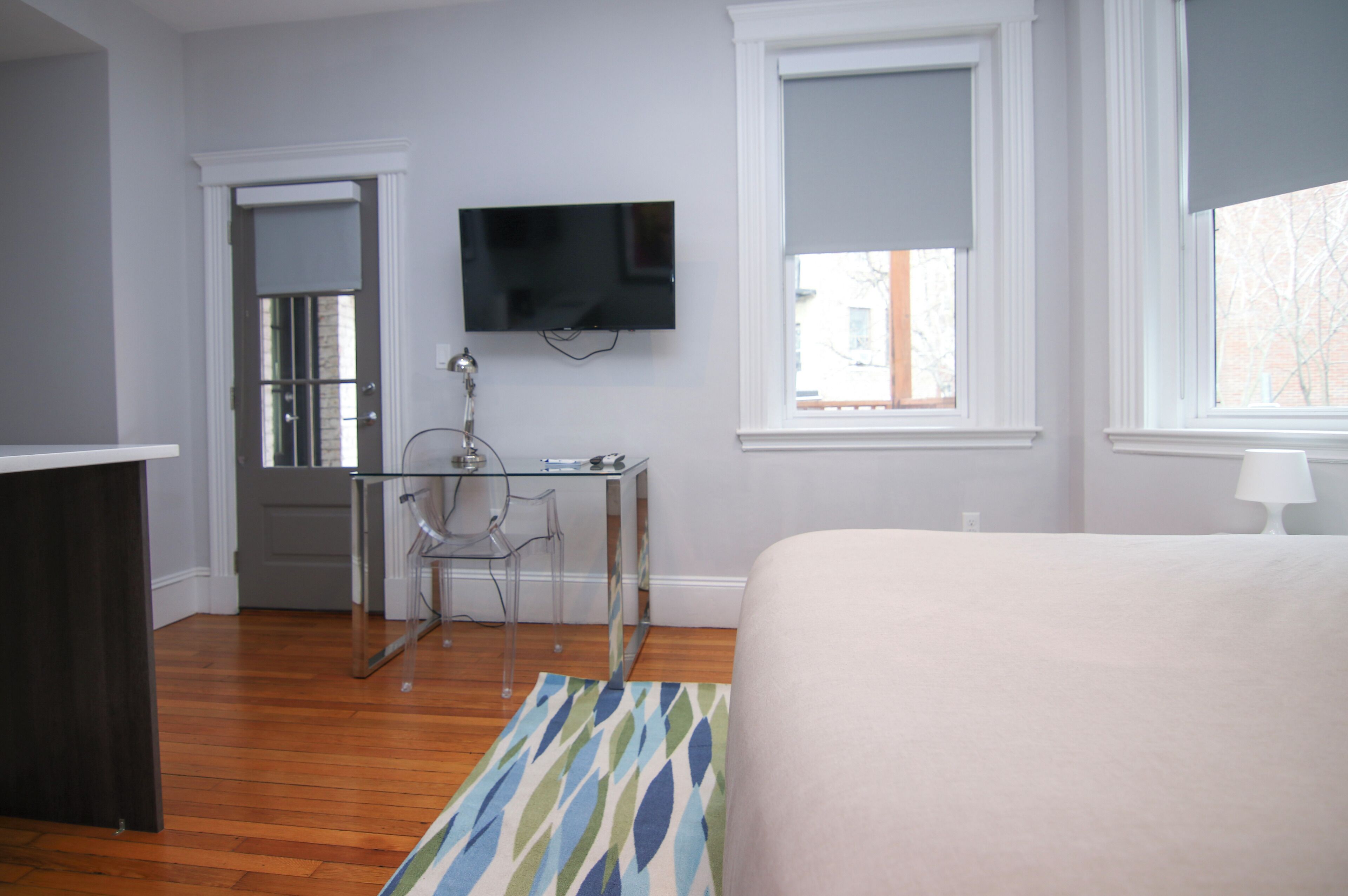 A bedroom with a bed, a TV mounted on the wall, a desk with a chair, and a window with a view of the outside.