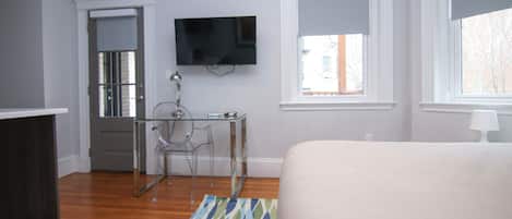 A bedroom with a bed, a TV mounted on the wall, a desk with a chair, and a window with a view of the outside.