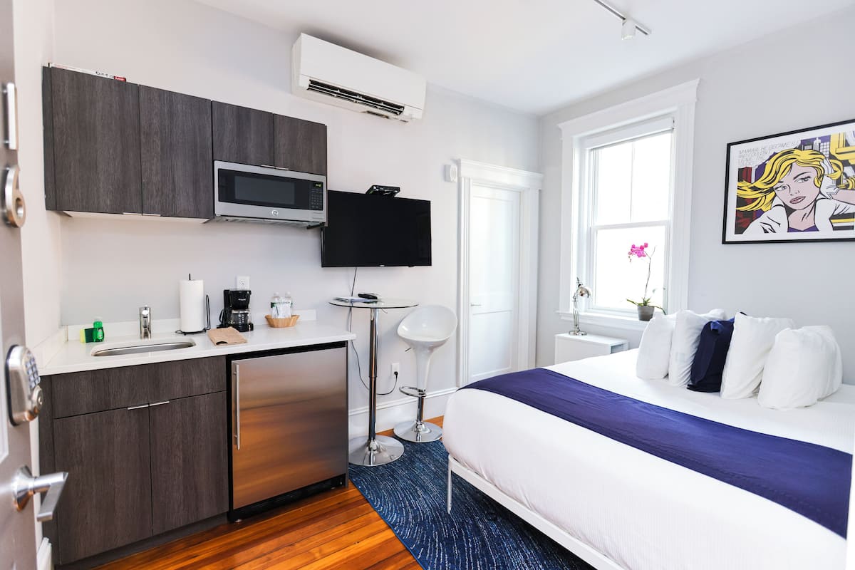 A modern hotel room with a kitchenette, a bed with a blue accent, a flat-screen TV, and a window with a view of the outdoors.