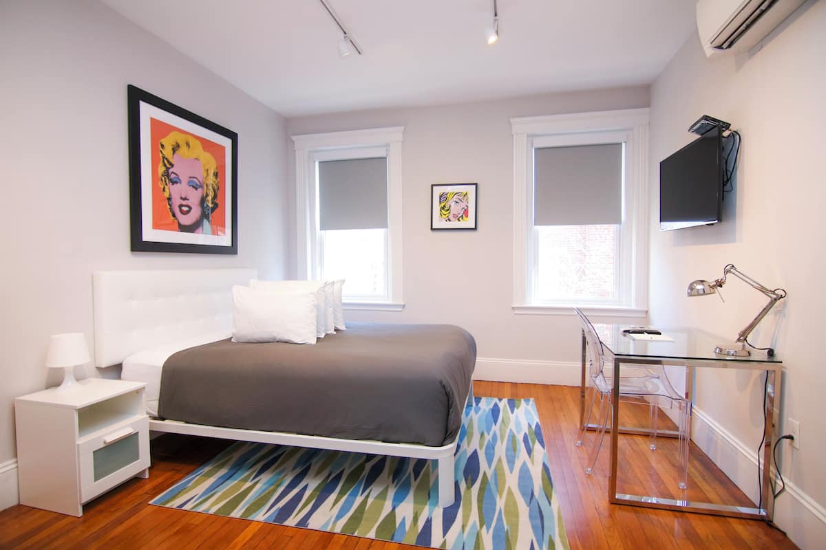 A modern bedroom with a bed, bedside table, lamp, and a desk with a chair.