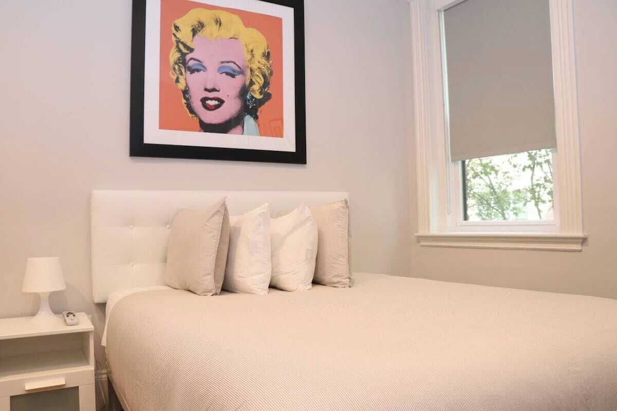 Een slaapkamer met een bed, kussens, een nachtkastje, een lamp en een lijst aan de muur.
