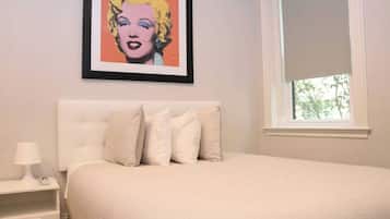 Een slaapkamer met een bed, kussens, een nachtkastje, een lamp en een lijst aan de muur.