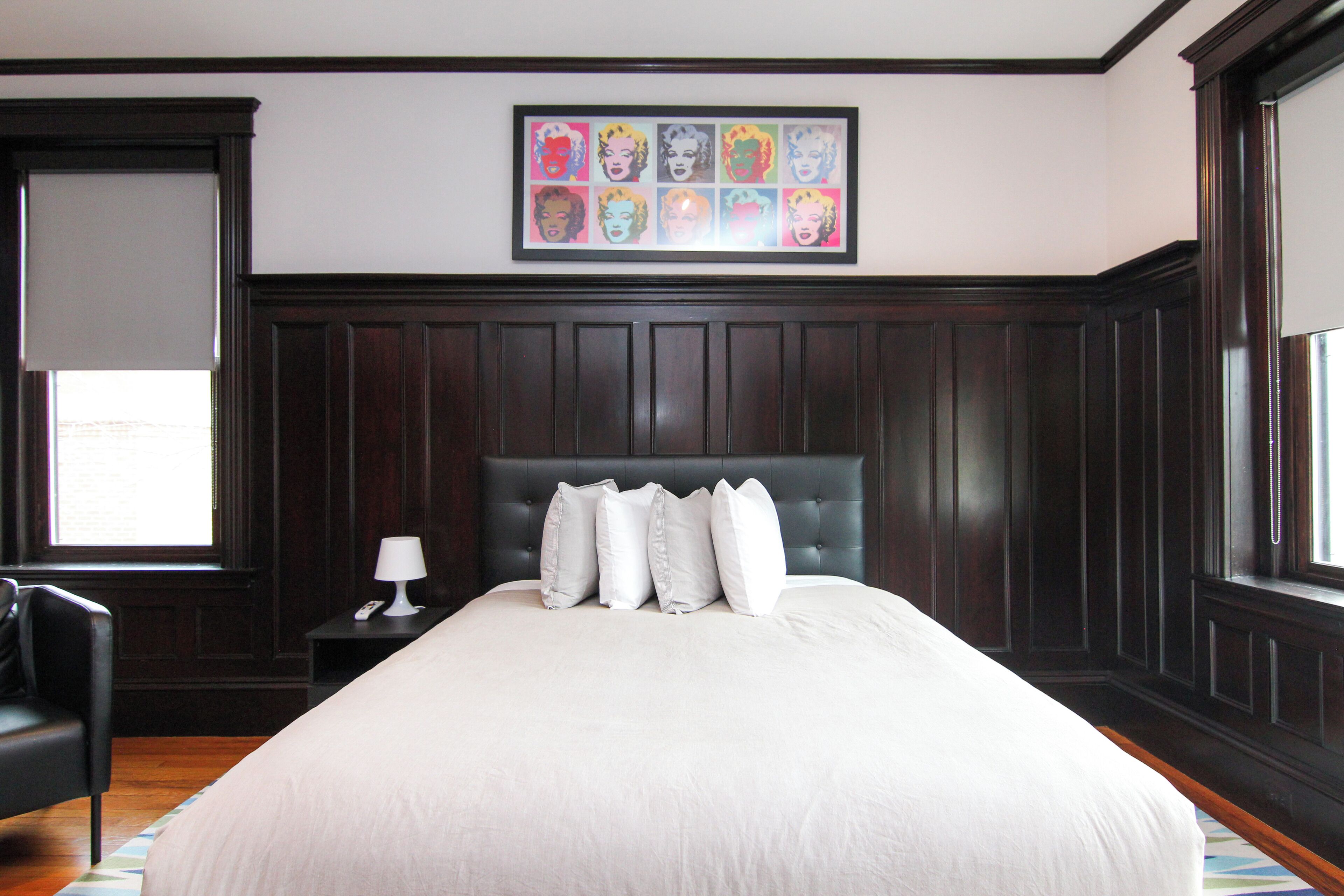 A hotel room with a large bed, dark wood paneling, a framed artwork above the bed, a bedside table with a lamp, and a window with a view.