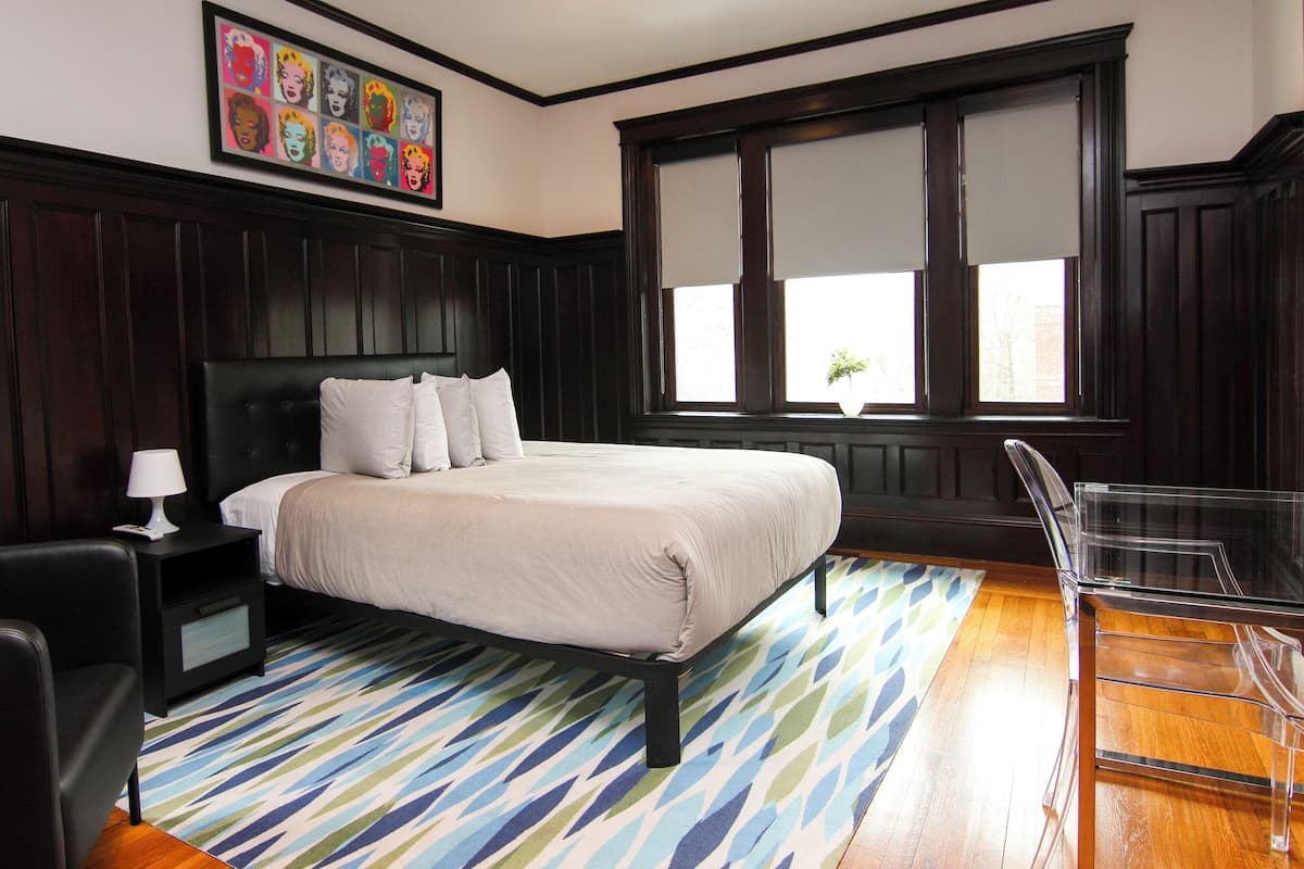 A bedroom with a bed, a nightstand, a chair, a window, and a wall with a framed picture.