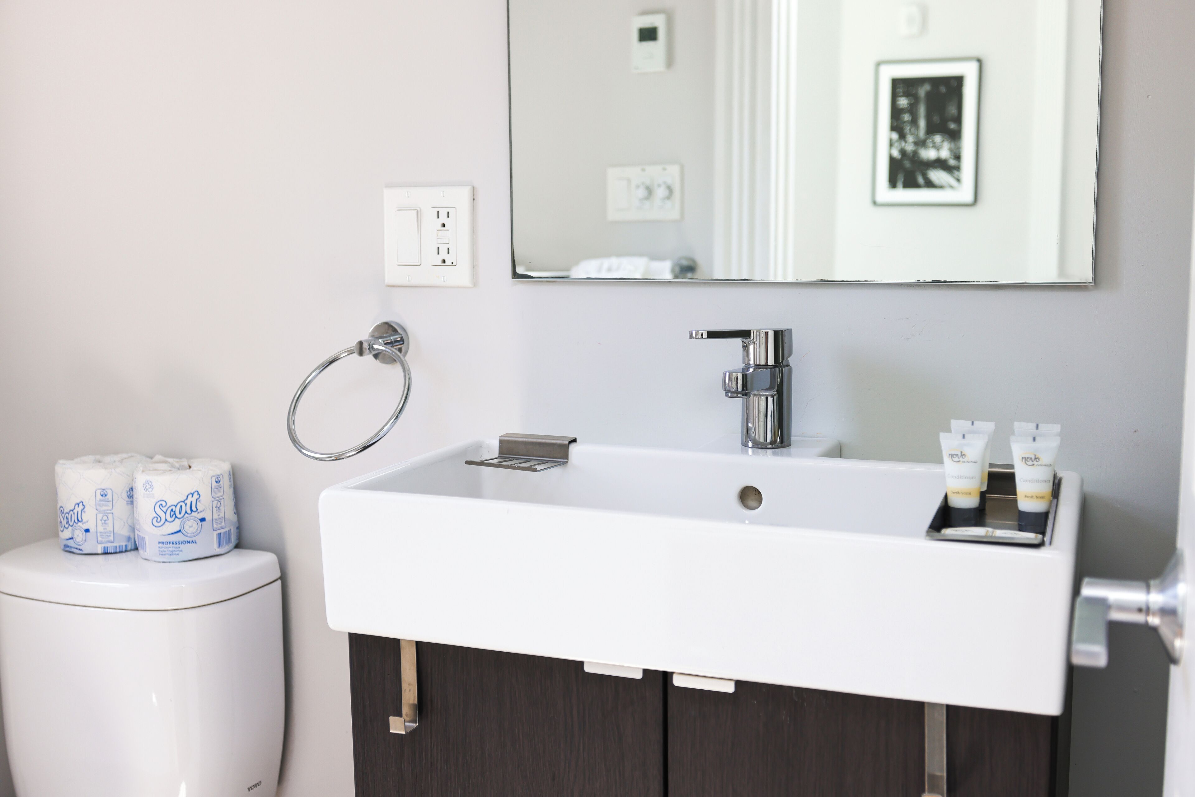 Ein Badezimmer mit weißem Waschbecken, dunklem Holzschrank und einem Spiegel über dem Waschbecken. Auf dem Waschbecken liegen zwei Toilettenpapierrollen und zwei Lotiontuben.