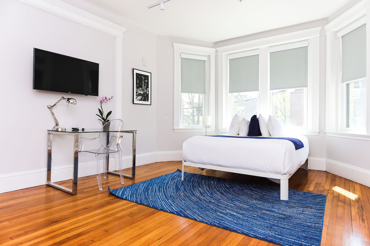 A bedroom with a bed, a desk with a chair, a TV, and large windows.