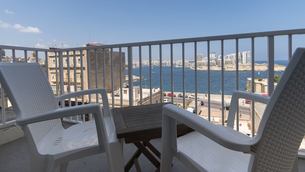 A balcony with white chairs and a small table, overlooking a coastal cityscape with buildings and a body of water. - Vallettastay Harbor Gem 2 bedroom  (Valletta)