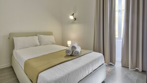 A neatly made bed with white linens and a beige throw, a wall-mounted light, and a window with curtains. - Vallettastay Harbor Gem 2 bedroom  (Valletta)