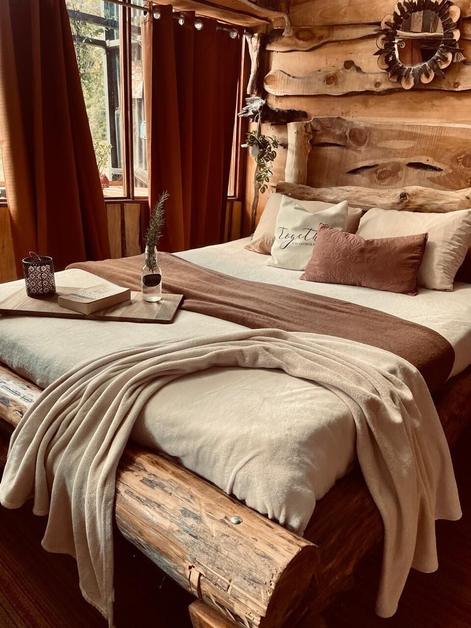 A wooden cabin interior with a bed, curtains, a wooden headboard, a bedside table with a book and a vase, and a window with curtains.
