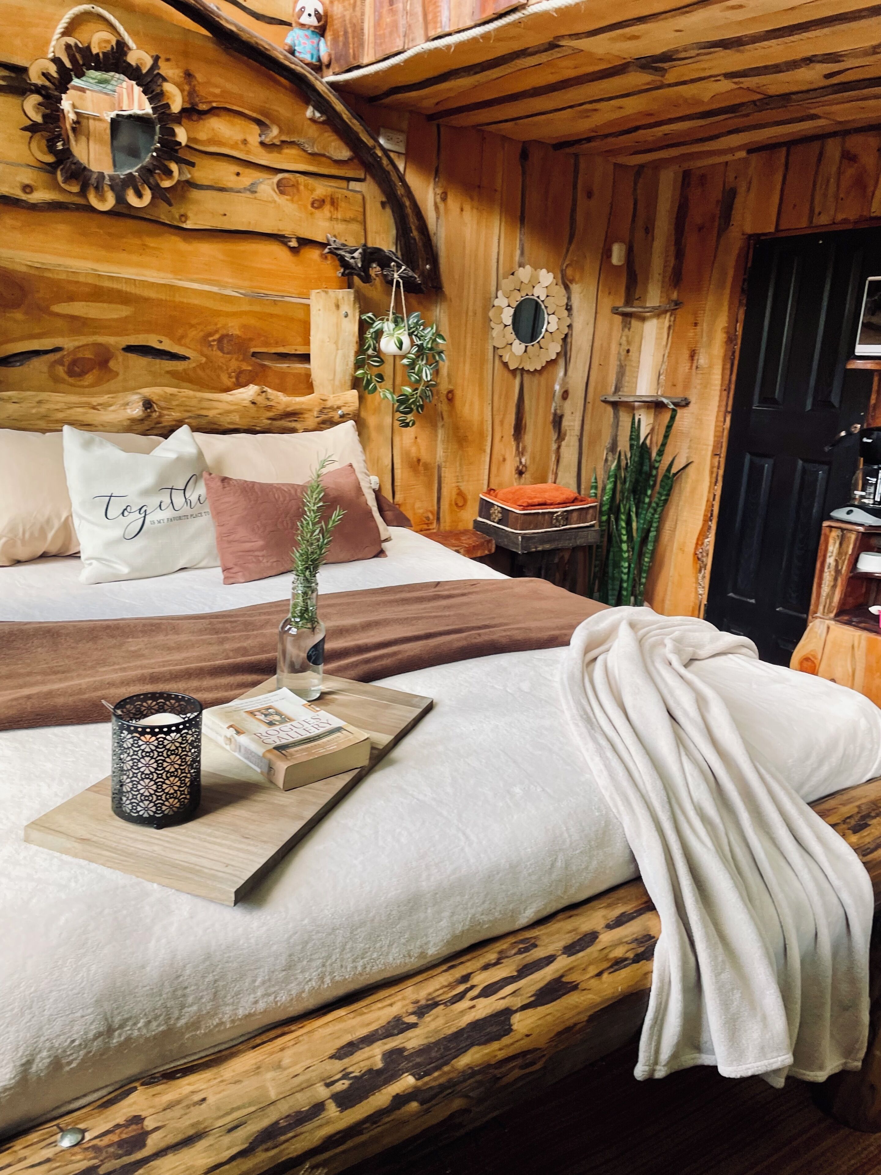 A cozy wooden cabin bedroom with a bed, pillows, a tray with a book and candle, and a small table with a plant.
