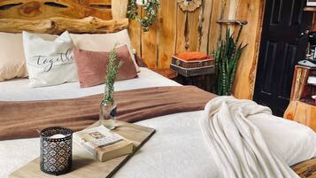A cozy wooden cabin bedroom with a bed, pillows, a tray with a book and candle, and a small table with a plant.