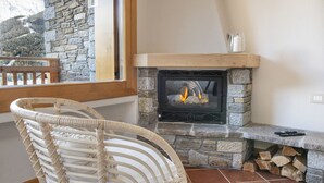 A stone fireplace with a fire, a wicker chair, and a view of a snowy landscape through a window. - InstaView - One Bedroom Apartment, Sleeps 4 (Pontagna)