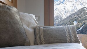 A bed with a white blanket and a grey pillow with a braided design, set against a mountain view. - InstaView - One Bedroom Apartment, Sleeps 4 (Pontagna)
