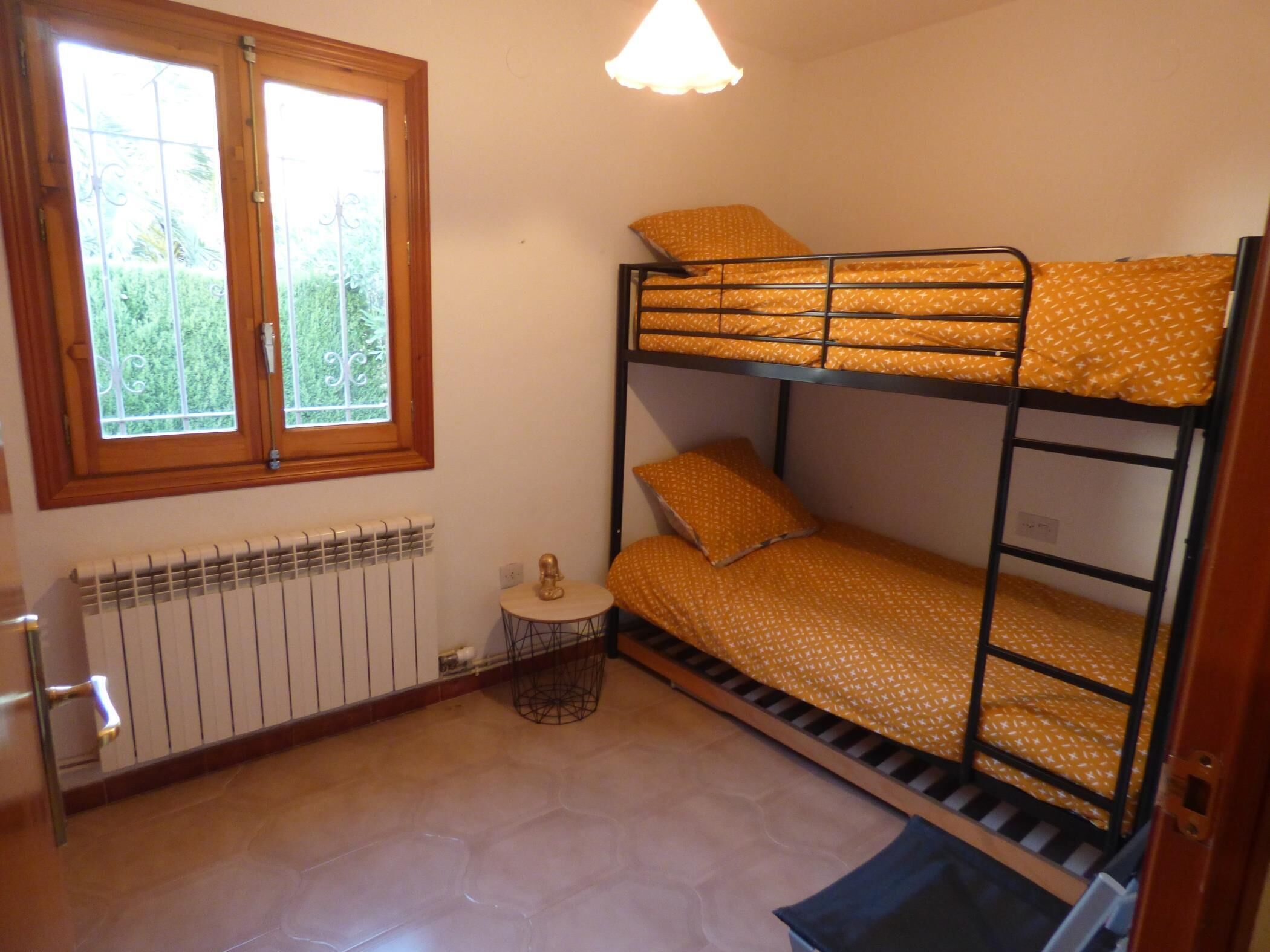 Habitación con literas, ventana, radiador y una mesita pequeña.