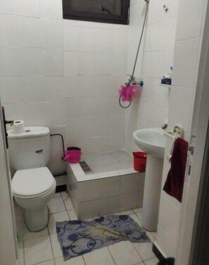 Un baño pequeño con inodoro, lavamanos y ducha. Hay dos cubetas, una rosa y una roja, y una alfombra azul estampada en el suelo.