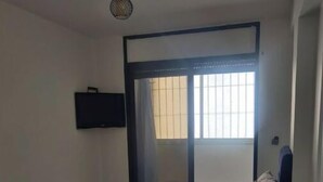 Een kamer met een bed, een bank, een salontafel, een aan de muur bevestigde televisie en een raam met zonwering.