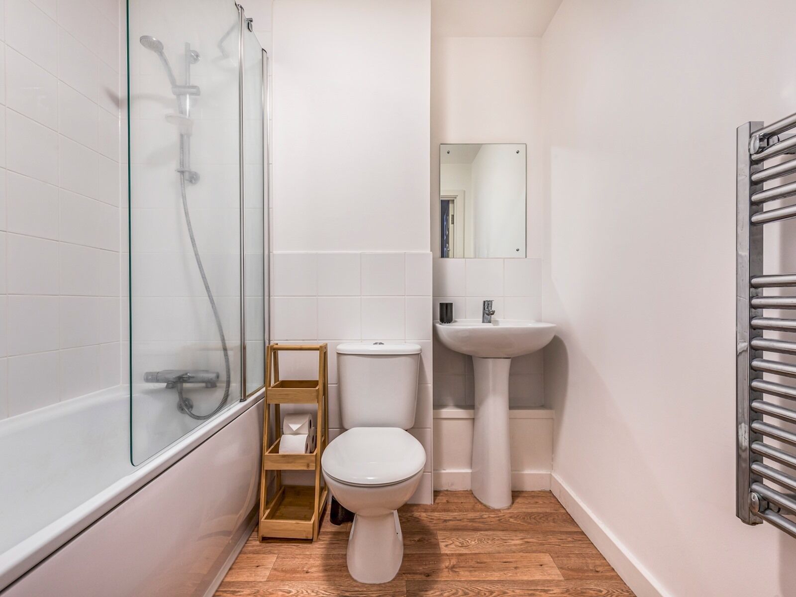 A modern bathroom with a glass shower enclosure, a toilet, a pedestal sink, and a wooden shelf.
