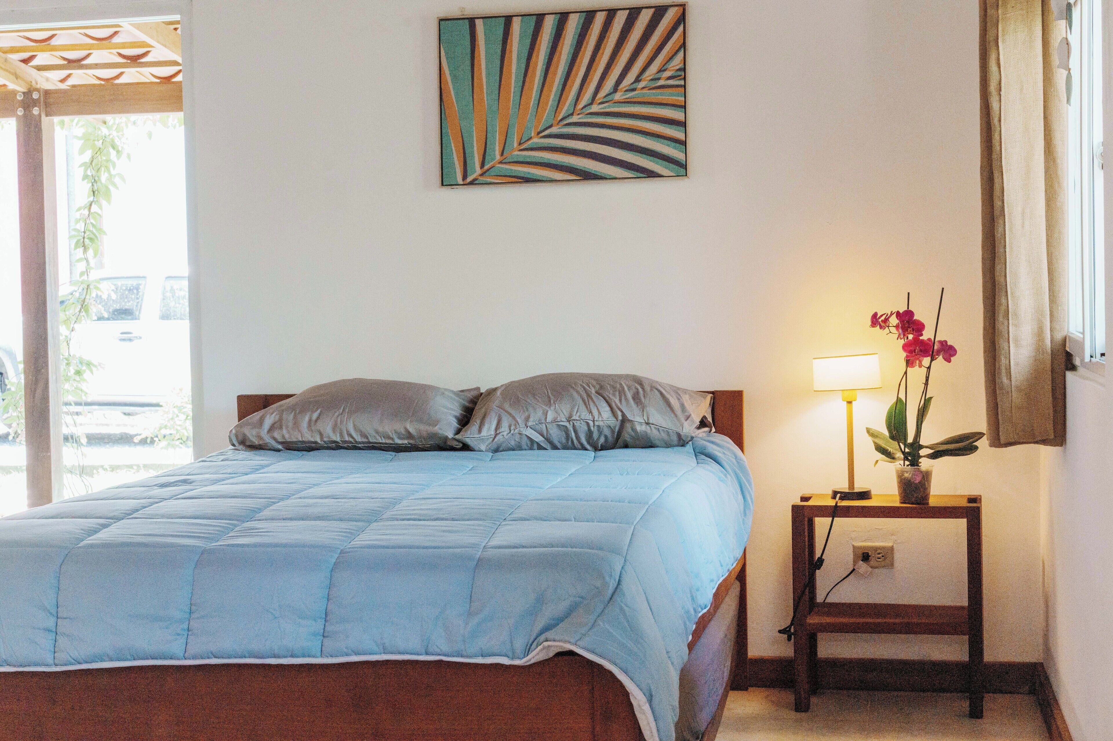 A bedroom with a bed, pillows, a nightstand with a lamp and a potted plant, and a wall art piece.