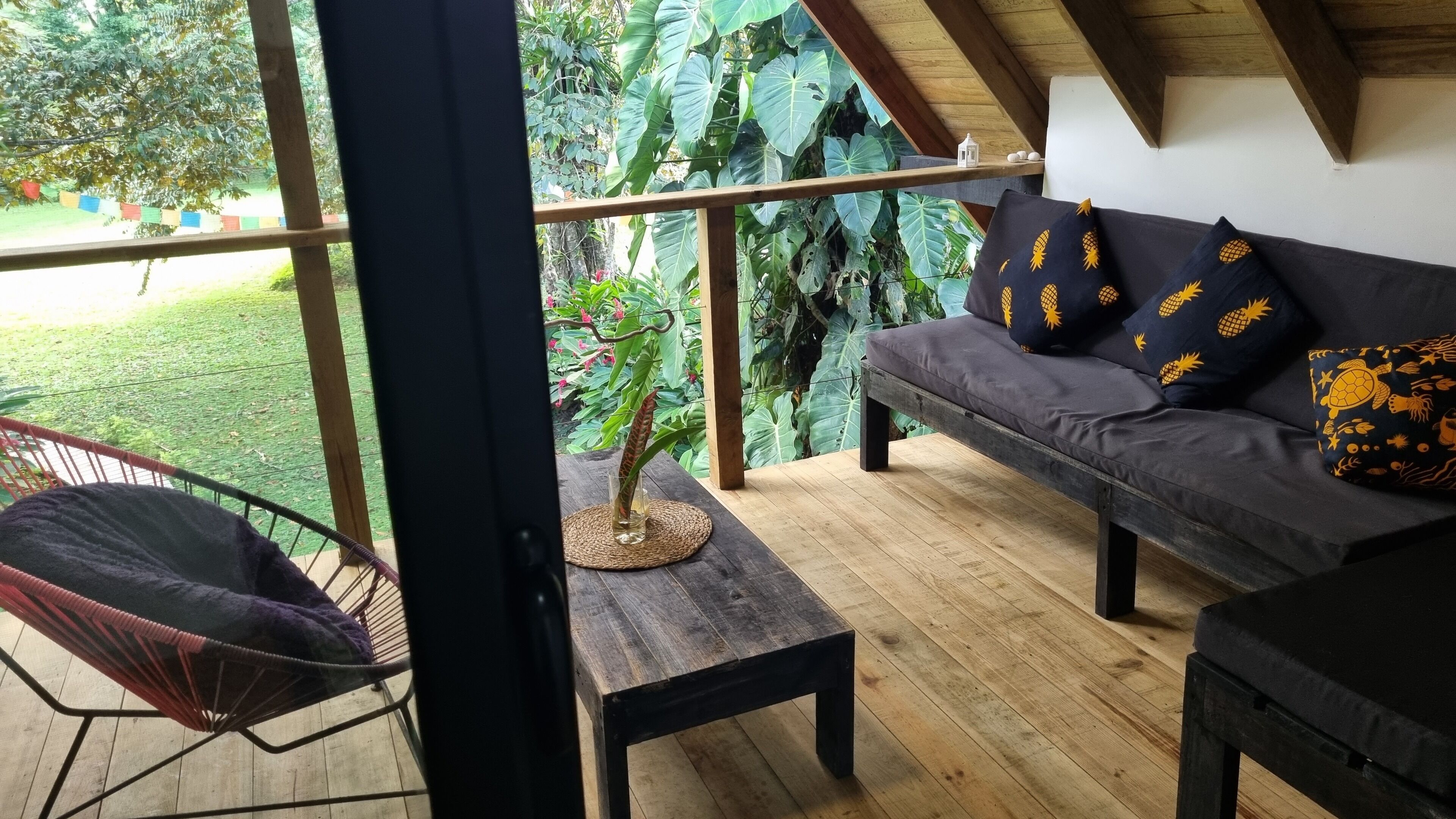 Ein gemütlicher Außenbereich mit Holztisch, gepolstertem Sessel und einer Couch mit tropischen Kissen.