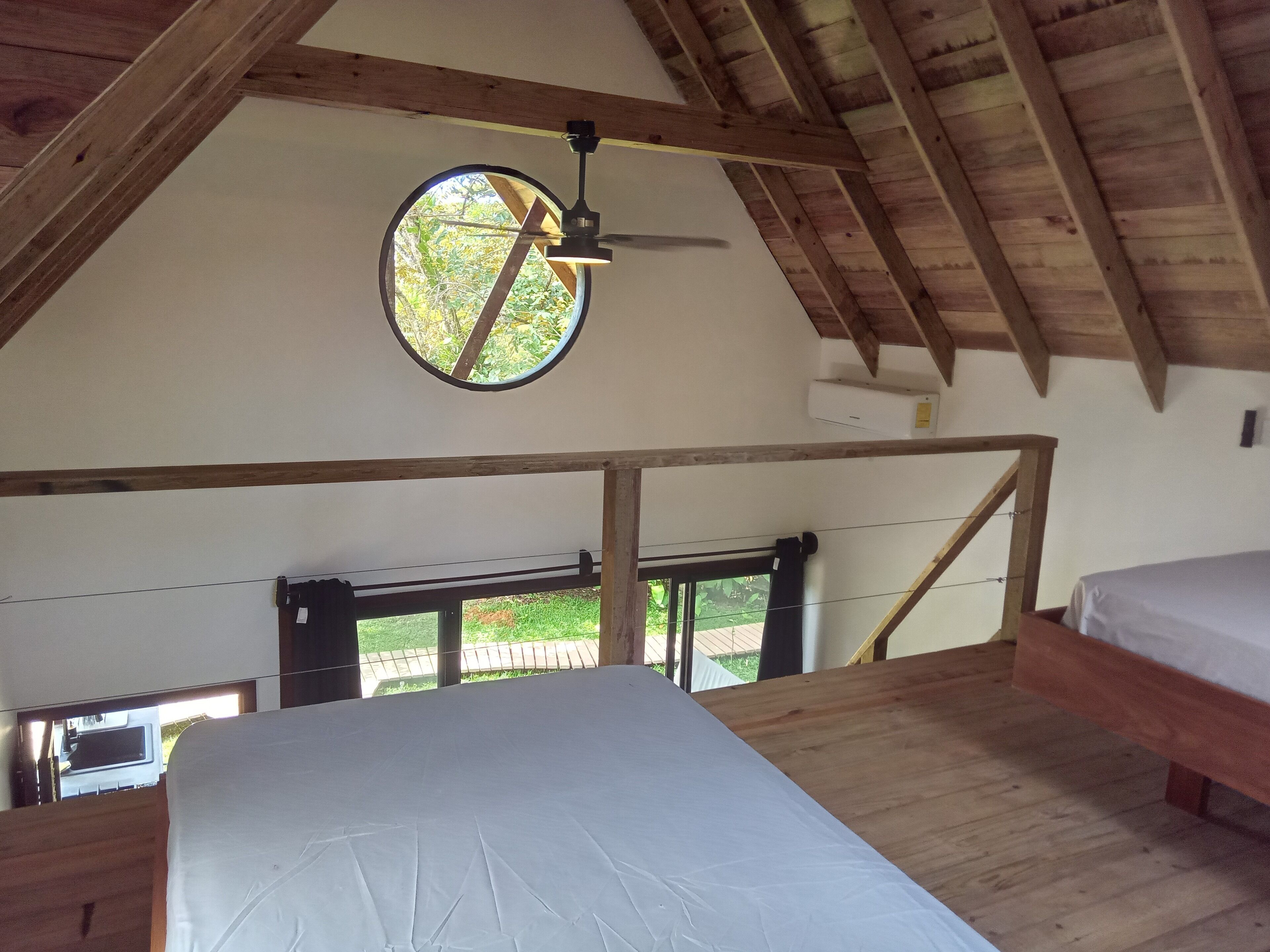 A high-ceilinged room with a wooden ceiling and a circular window. A bed is positioned against the wall, and a ceiling fan is visible.