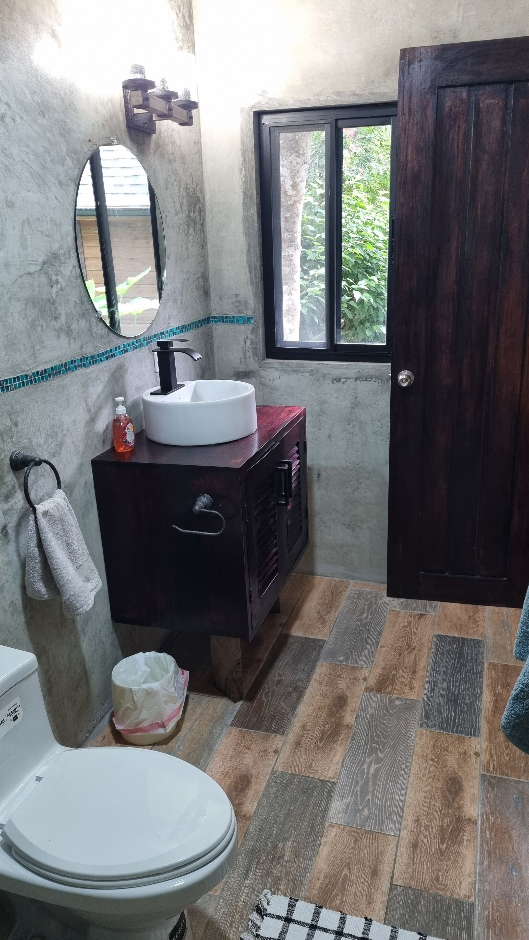 A bathroom with a white toilet, a wooden vanity with a white sink, a mirror, and a window with a view of greenery.